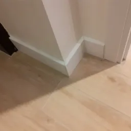 Szczegółowe ujęcie wykończenia podłogi z jasnych płytek imitujących drewno, z białymi listwami przypodłogowymi i narożnikiem przy ościeżnicy drzwi.