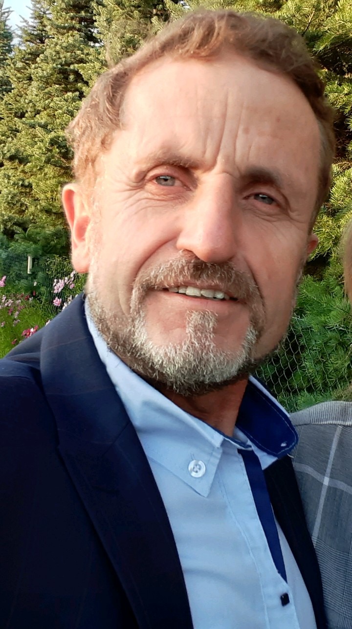 Portret uśmiechniętego mężczyzny w niebieskiej koszuli i marynarce na tle zieleni i kwiatów.