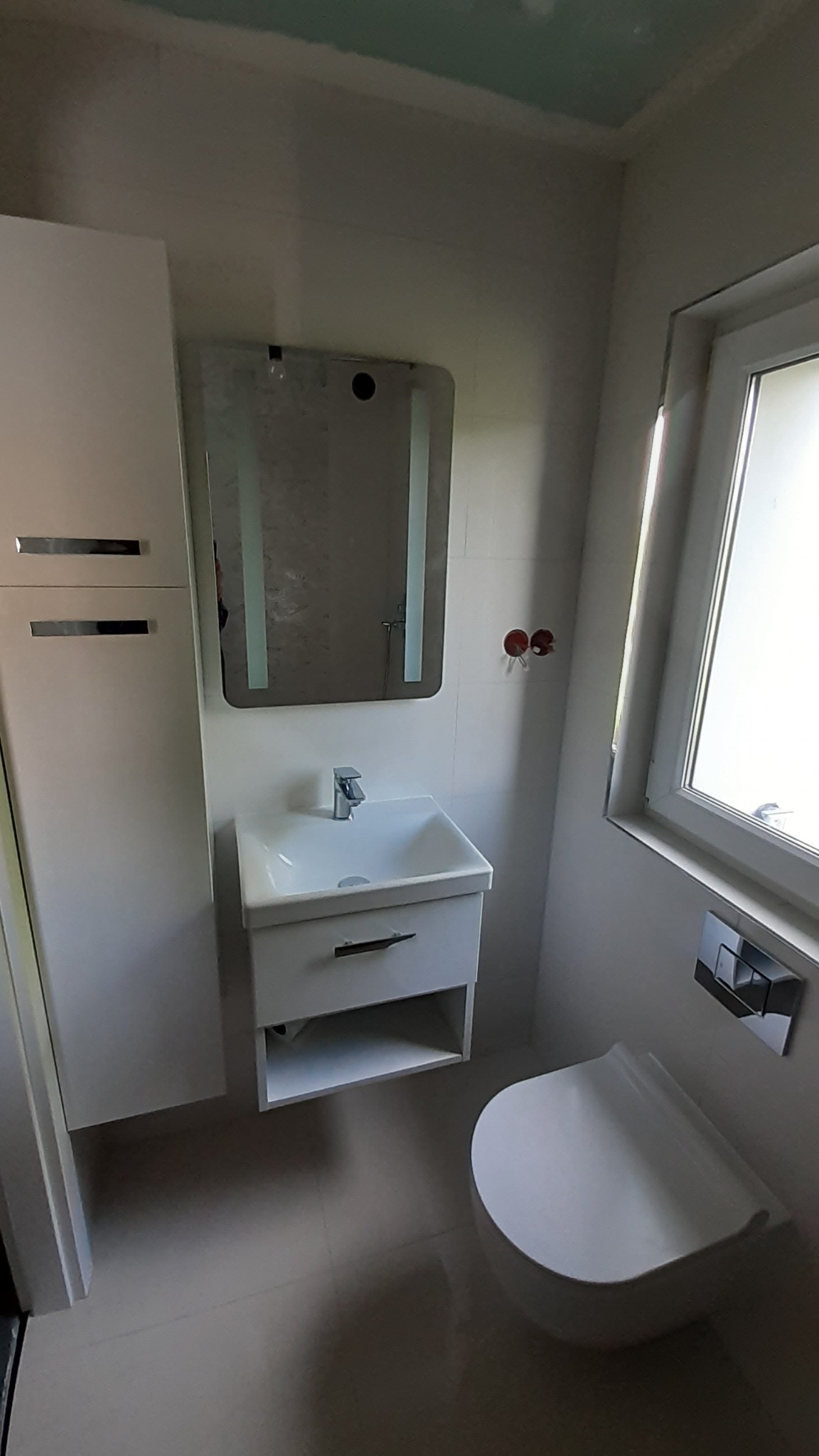 Nowoczesna, jasna łazienka z białymi meblami: szafka z umywalką, lustro z podświetleniem LED, wisząca toaleta. Widoczne niedokończone wykończenie sufitu i wystające kable elektryczne.