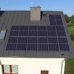 Pelsun - Panele słoneczne na szarym dachu domu jednorodzinnego z oknami dachowymi i kominami, widok z góry na zadbany ogród i drugi dom z panelami w tle.