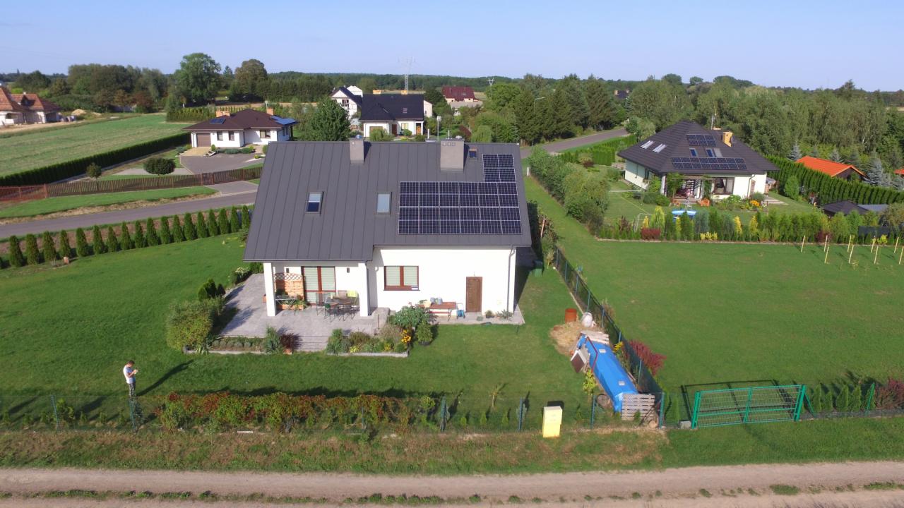 Dom jednorodzinny z szarym dachem i panelami słonecznymi, widok z lotu ptaka, w tle inne domy i zieleń.