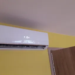 Biały klimatyzator Sinclair zamontowany na żółtej ścianie, widoczny fragment drewnianej ościeżnicy drzwi