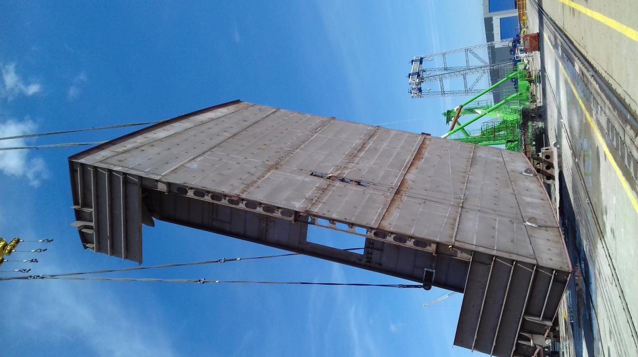 Metalowa konstrukcja hali podnoszona przez dźwig na tle błękitnego nieba. Widoczne spawane łączenia blach i otwory w konstrukcji.