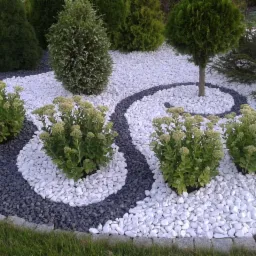 Uporządkowany ogród z krzewami, drzewami i wijącymi się ścieżkami z białych i szarych kamyków, oddzielonych obrzeżem z kostki brukowej.