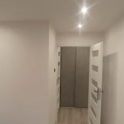 Wnętrze po remoncie: Świeżo pomalowane ściany w odcieniach bieli, sufit z trzema punktowymi lampami LED, otwarte białe drzwi z przeszkleniami prowadzące do pomieszczenia z szarą szafą przesuwną...