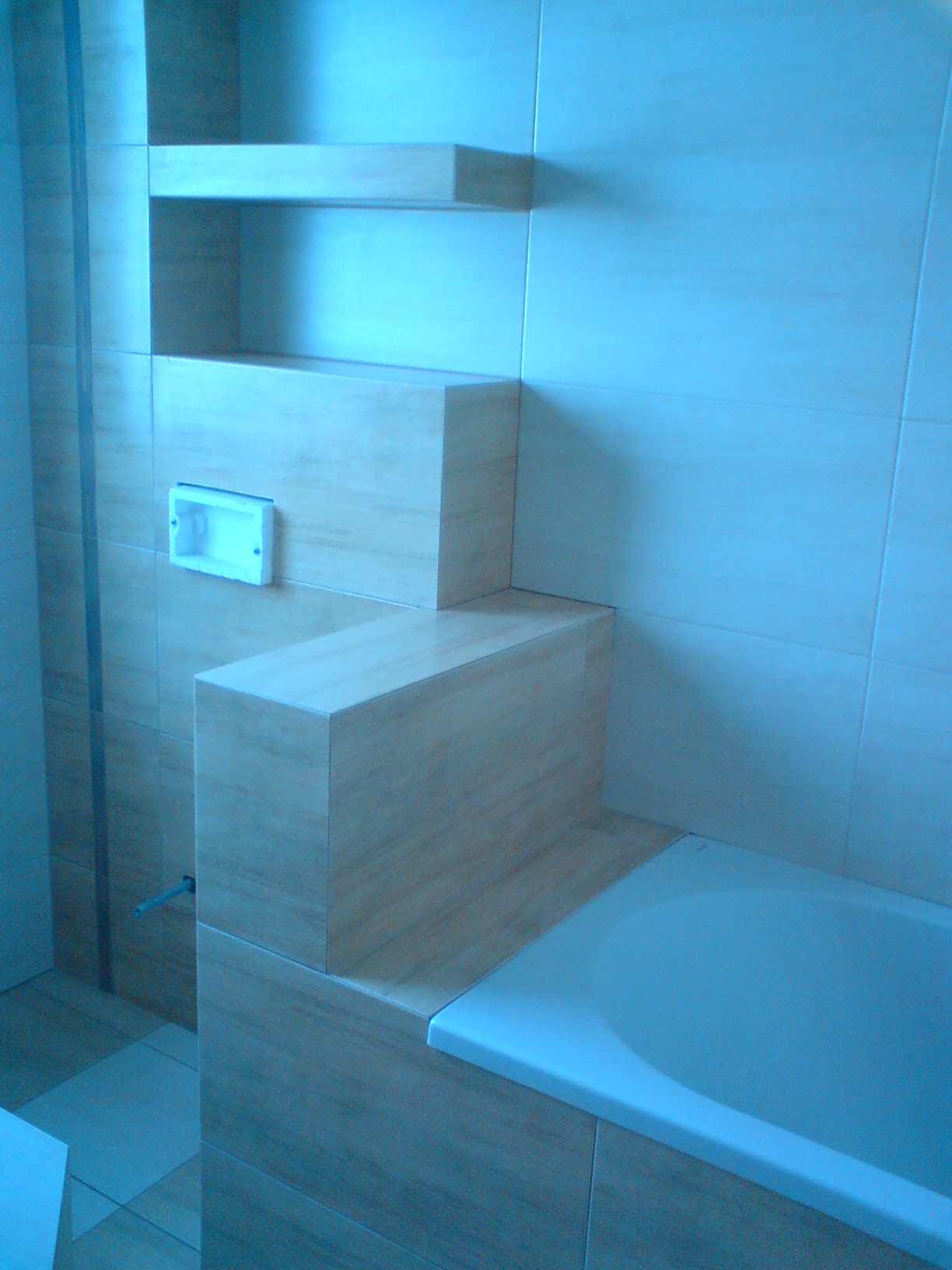 Wykończona łazienka z wanną, półkami i zabudową z płytek ceramicznych imitujących drewno, widoczna puszka elektryczna.