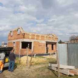 Budowa domu jednorodzinnego w trakcie murowania ścian z pustaków ceramicznych; widoczny murarz w oknie, samochód z otwartym bagażnikiem i dwoma osobami w pomarańczowych spodniach oraz leżące...