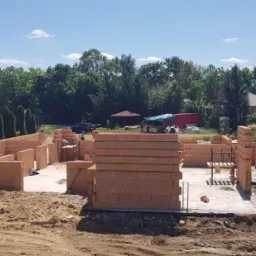 Wznoszenie ścian parteru z bloczków ceramicznych na fundamencie, widoczny transport bloczków na budowę, w tle zieleń drzew i zabudowa jednorodzinna.