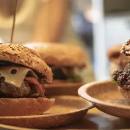Dwa burgery z wołowiną, boczkiem i serem na biodegradowalnych talerzach, jeden z wbitym patyczkiem, w tle rozmyte postacie i kolejne burgery.