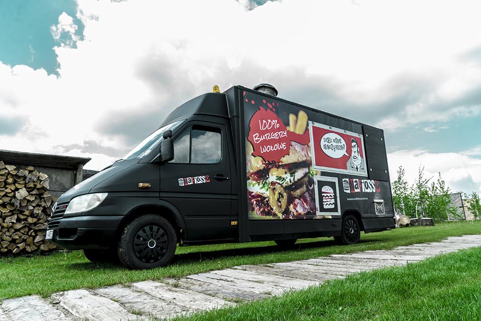 Czarny food truck z reklamą burgerów na tle zielonej trawy i sterty drewna.