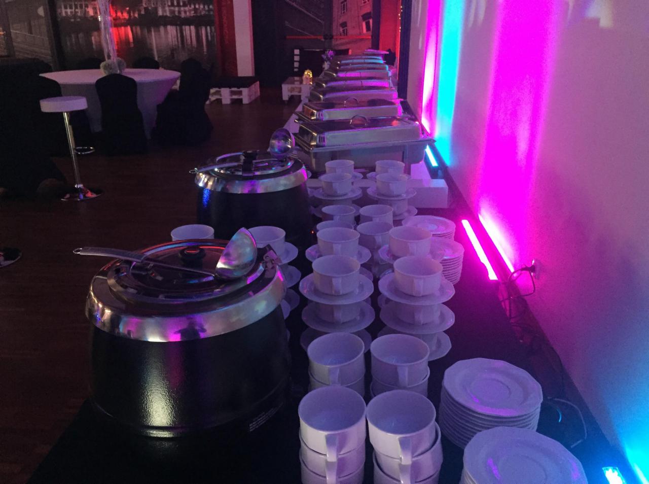Przygotowany bufet z podgrzewaczami do zup i stosami białych filiżanek na spodkach, oświetlony neonowym, niebiesko-różowym światłem, w tle stoliki z białymi obrusami i dekoracją.