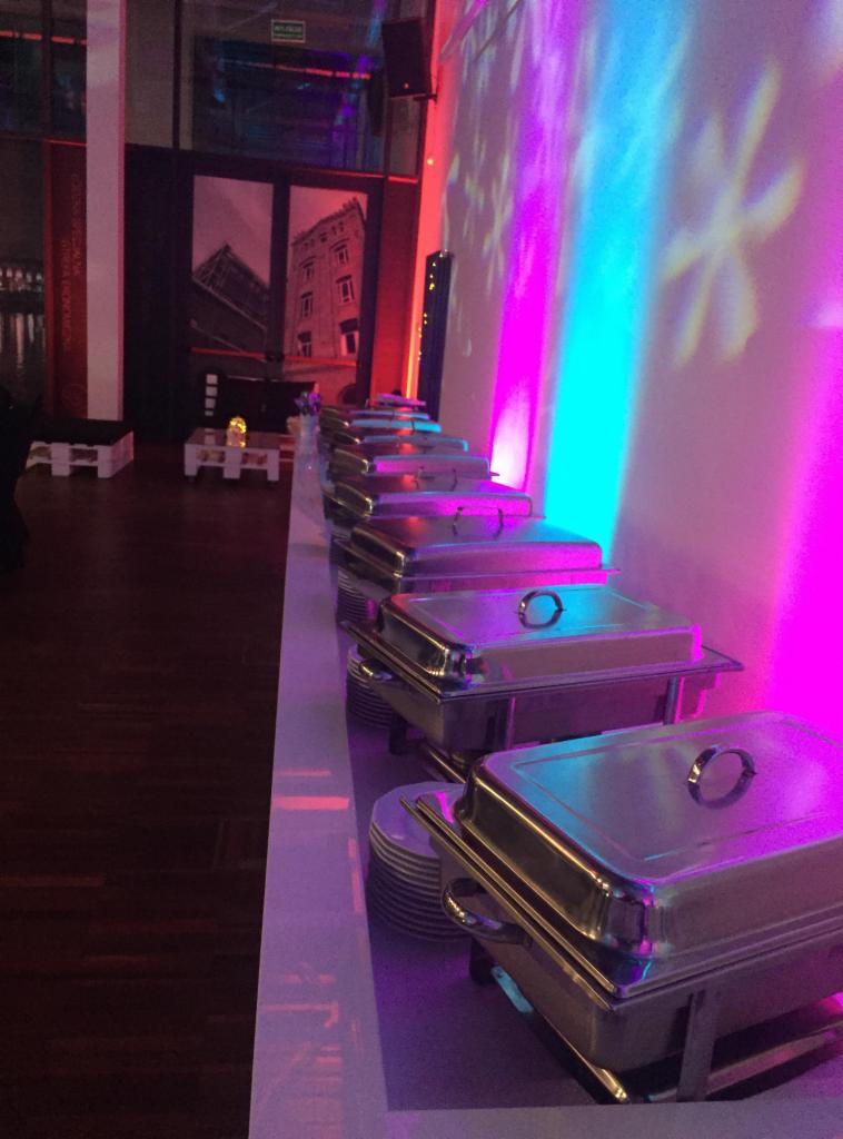 Rząd metalowych bemarów z potrawami ustawionych na stole podczas imprezy, oświetlonych niebiesko-różowym światłem, z widocznymi talerzami obok, w tle dekoracja z motywem miejskim.