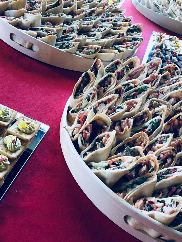 Tortilla wrap przekrojone na pół, ułożone rzędami na drewnianej tacy, obok małe kanapeczki na srebrnej tacy, całość na czerwonym obrusie.