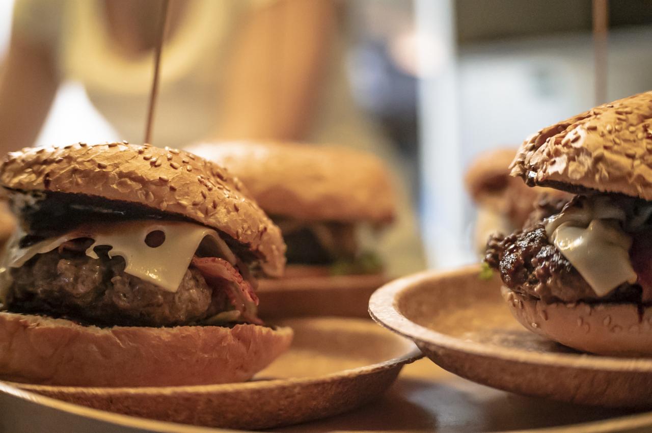 Dwa burgery z wołowiną, boczkiem i serem na biodegradowalnych talerzach, jeden z wbitym patyczkiem, w tle rozmyte postacie i kolejne burgery.