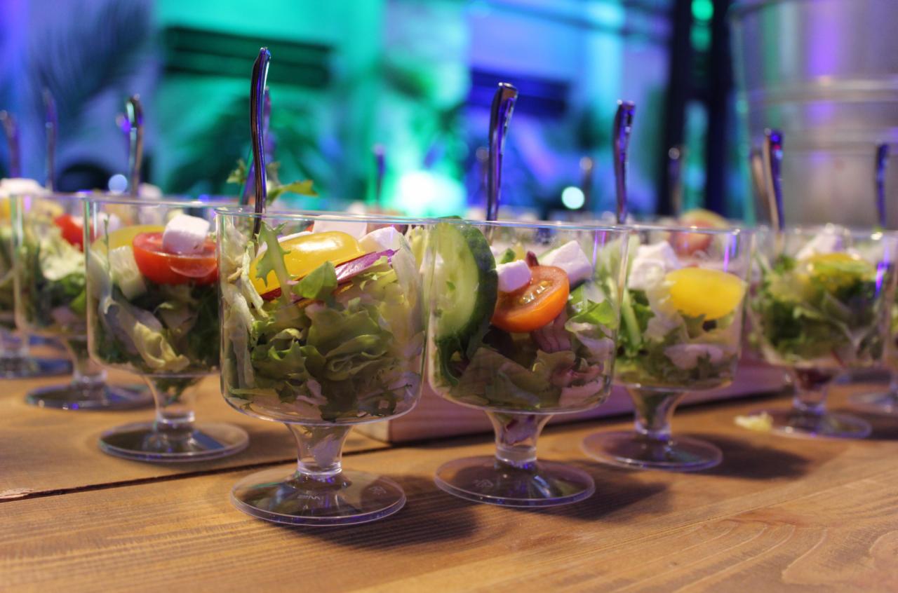 Szereg szklanych pucharków na krótkich nóżkach wypełnionych sałatką ze świeżych warzyw, pomidorków koktajlowych, kostek fety i zwieńczonych srebrnymi łyżeczkami, ustawionych na drewnianym blacie,...