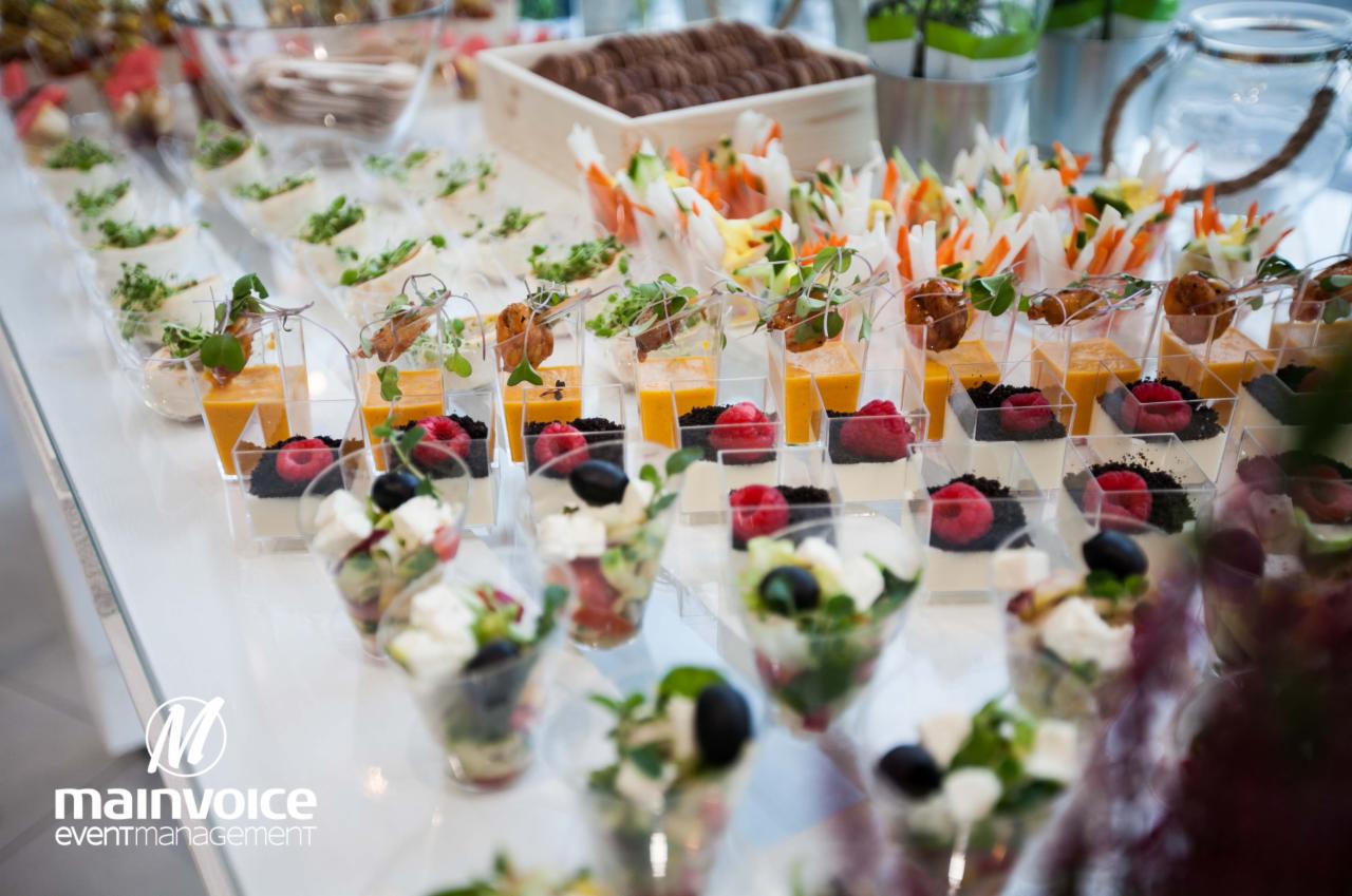 Różnorodne przekąski w małych przezroczystych naczyniach na białym stole, w tym sałatki z fetą i oliwkami, desery z malinami i czarnym kruszonem, krem z dyni z dekoracją, oraz spring rollsy...