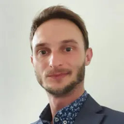 Portret mężczyzny w niebieskiej marynarce i koszuli w kwiaty na jasnym tle. Uśmiechnięty, z krótką brodą i starannie uczesanymi włosami. Dobre oświetlenie, wyraźne rysy twarzy.