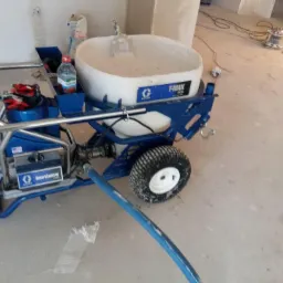 Agregat hydrodynamiczny Graco T-MAX 506 z pełnym zbiornikiem gotowej masy szpachlowej, butelką wody i rękawicami roboczymi, przygotowany do aplikacji w surowym wnętrzu.