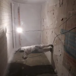 Pracownik w kombinezonie ochronnym natryskowo aplikuje białą substancję na ścianę piwnicy, oświetloną jasną żarówką; widoczne ślady wilgoci i uszkodzeń na ścianach.