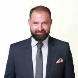 Portret uśmiechniętego mężczyzny z brodą w ciemnym garniturze i szarym krawacie, z beżową poszetką w kieszonce.