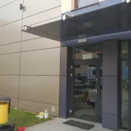 Nowoczesne wejście do budynku z zadaszeniem szklanym, widoczne puszki farby i wiadro sugerujące świeże malowanie elewacji.