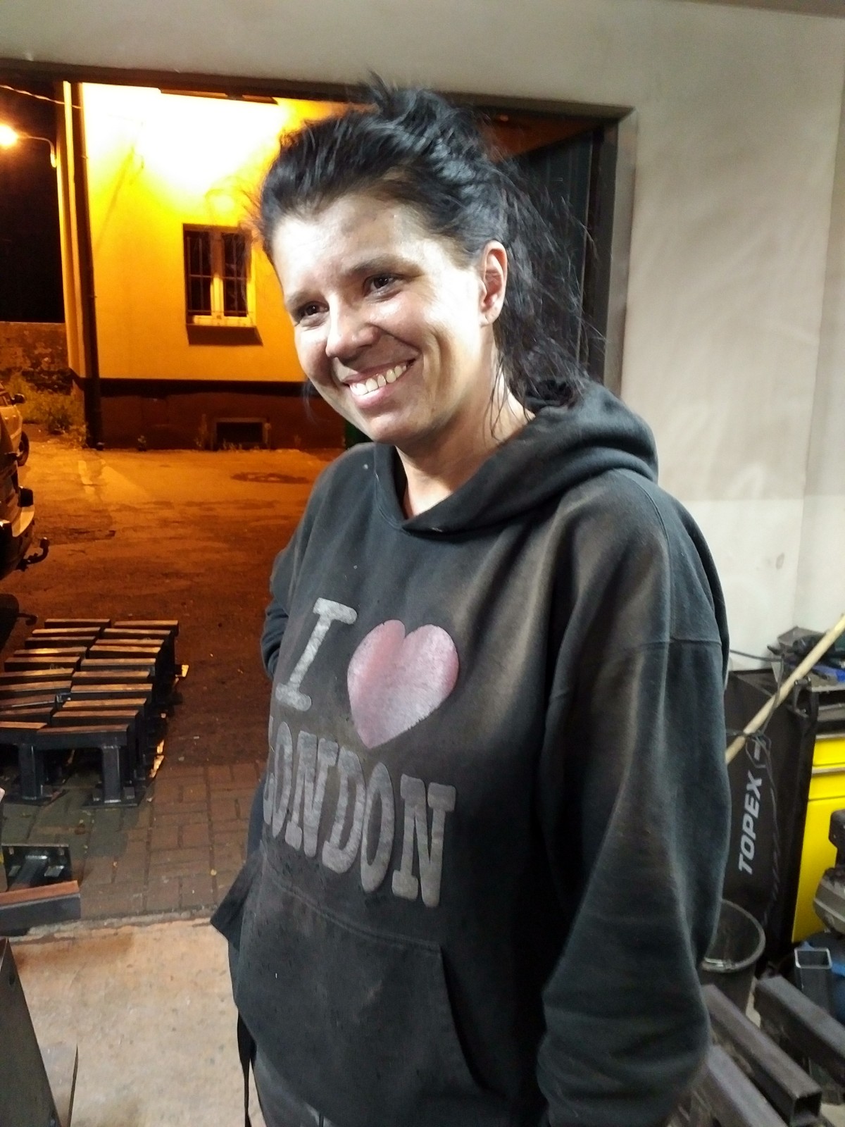 Uśmiechnięta kobieta w bluzie z napisem 'I love London' stoi w warsztacie, w tle widoczny fragment budynku z oknem i metalowe elementy konstrukcyjne.