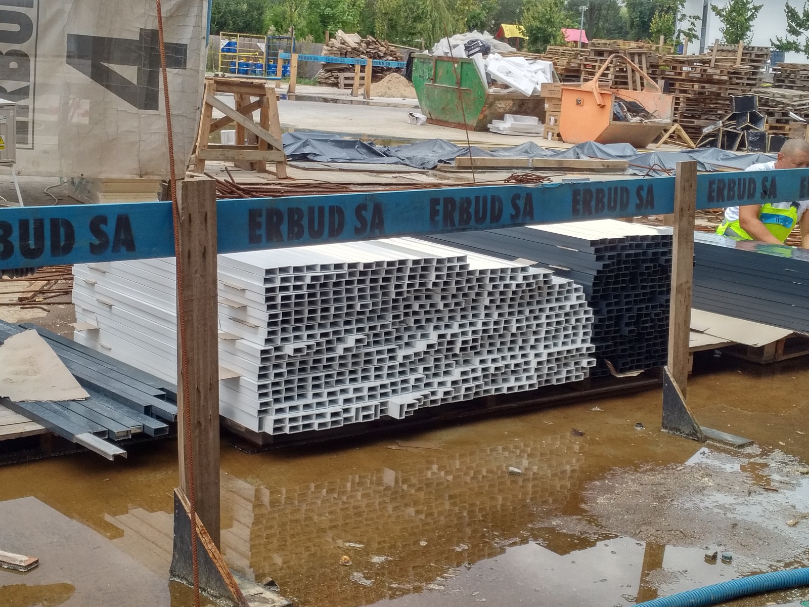 Stosy białych i czarnych profili metalowych o przekroju kwadratowym, składowane na paletach w błotnistej wodzie na placu budowy, z widocznym logo Erbud SA na belkach zabezpieczających.