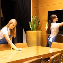BLUE - MART - Dwie kobiety w uniformach sprzątają nowoczesną salę konferencyjną: jedna wyciera duży drewniany stół, druga odkurza telewizor kolorową miotełką.