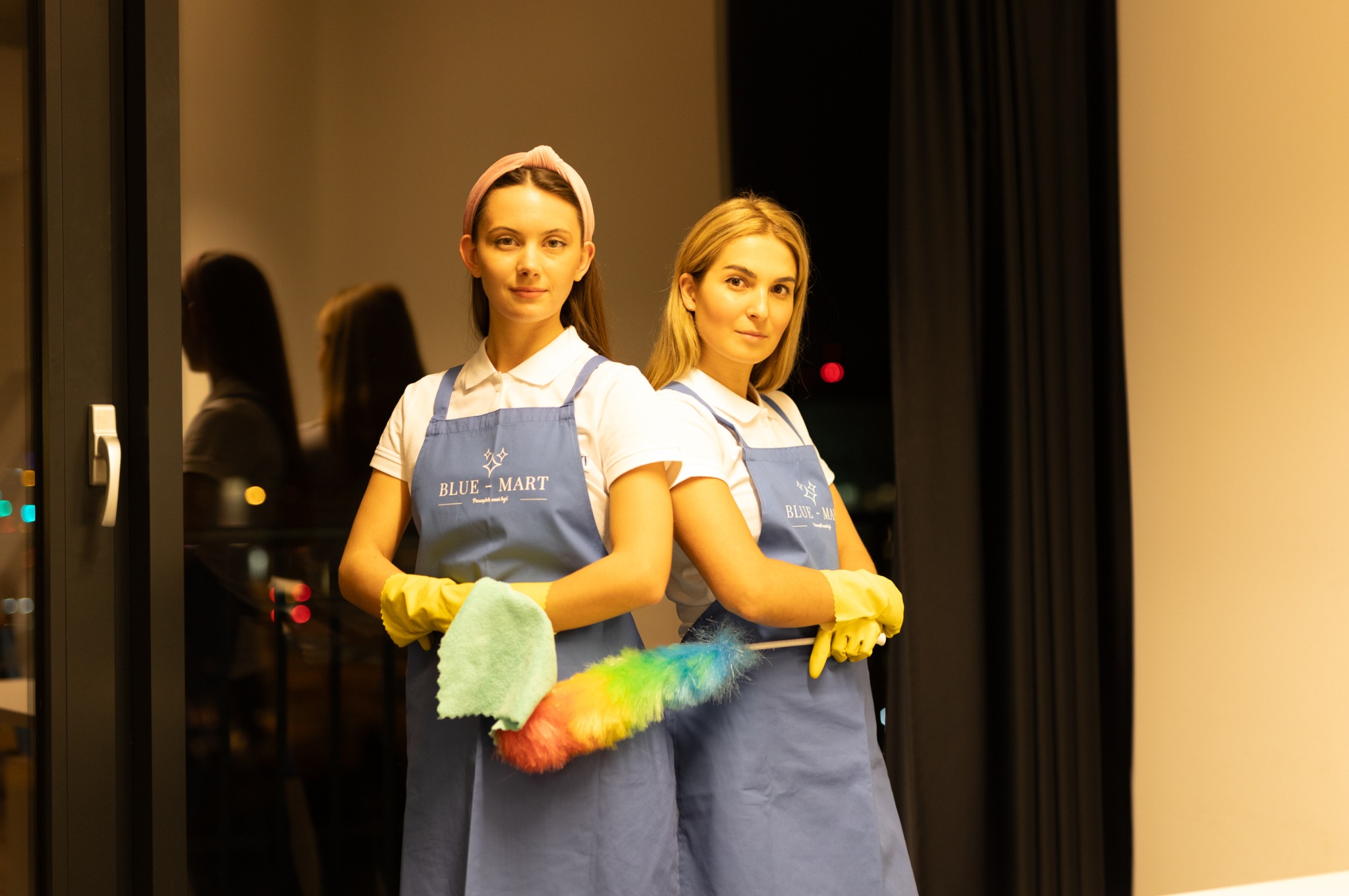 Dwie uśmiechnięte kobiety w fartuchach Blue-Mart, w żółtych rękawicach trzymające ściereczkę i kolorową miotełkę do kurzu, na tle okna i zasłon.