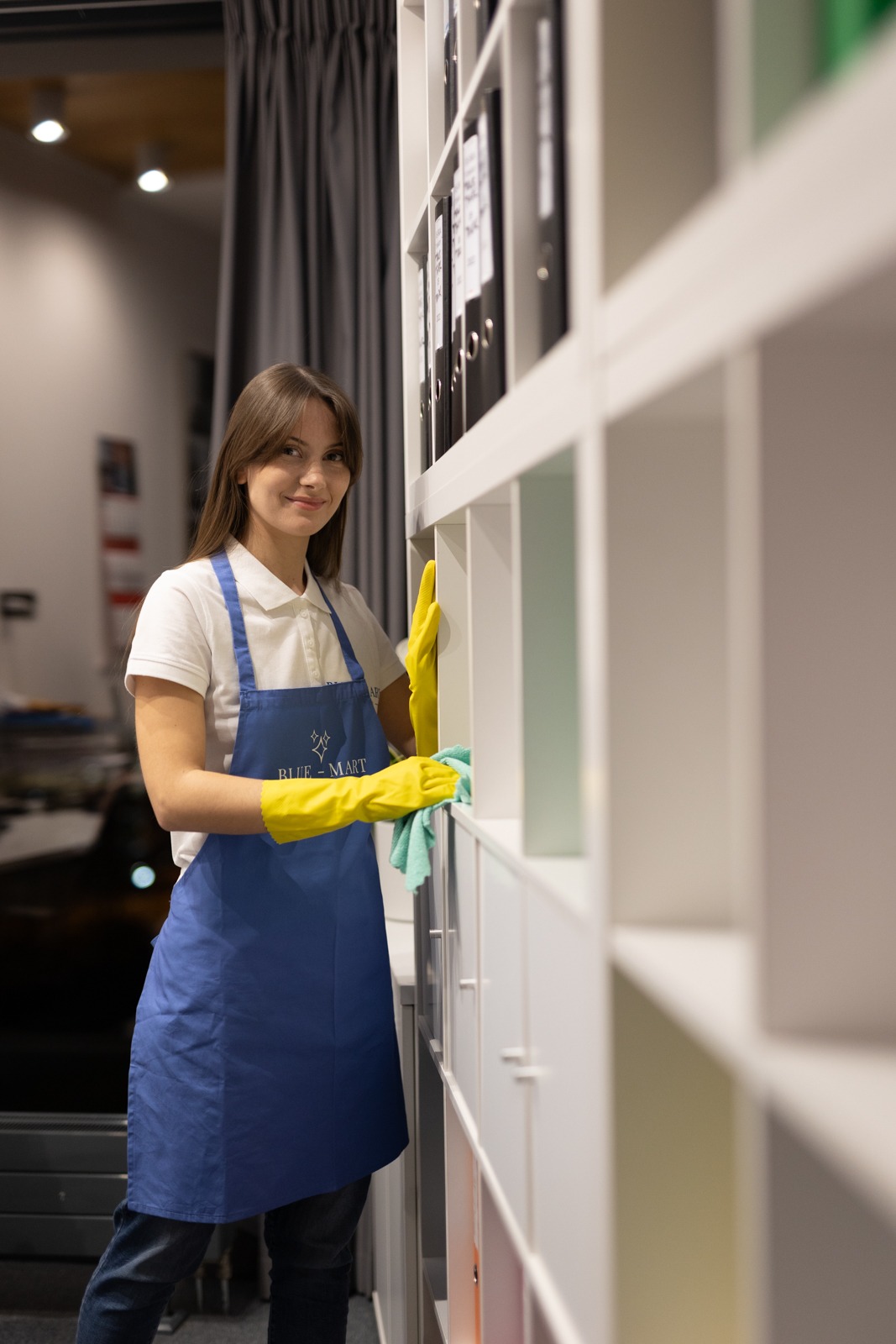 Pracownica w żółtych rękawicach i niebieskim fartuchu czyści półki regału w biurze, używając ściereczki. Widoczne segregatory i elementy wyposażenia.