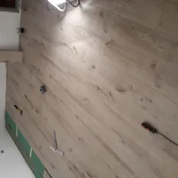 Montaż paneli podłogowych w trakcie realizacji, widoczny fragment ściany z listwą przypodłogową, poziomica, młotek, śrubokręt i paczka paneli.