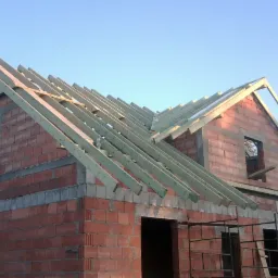 Surowy stan domu murowanego z widoczną konstrukcją więźby dachowej i niezadaszonymi otworami okiennymi, w tle błękitne niebo.