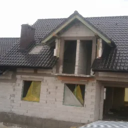 Dom murowany w trakcie budowy, widoczna ciemna dachówka, komin oraz okna zabezpieczone folią, surowy stan budynku.