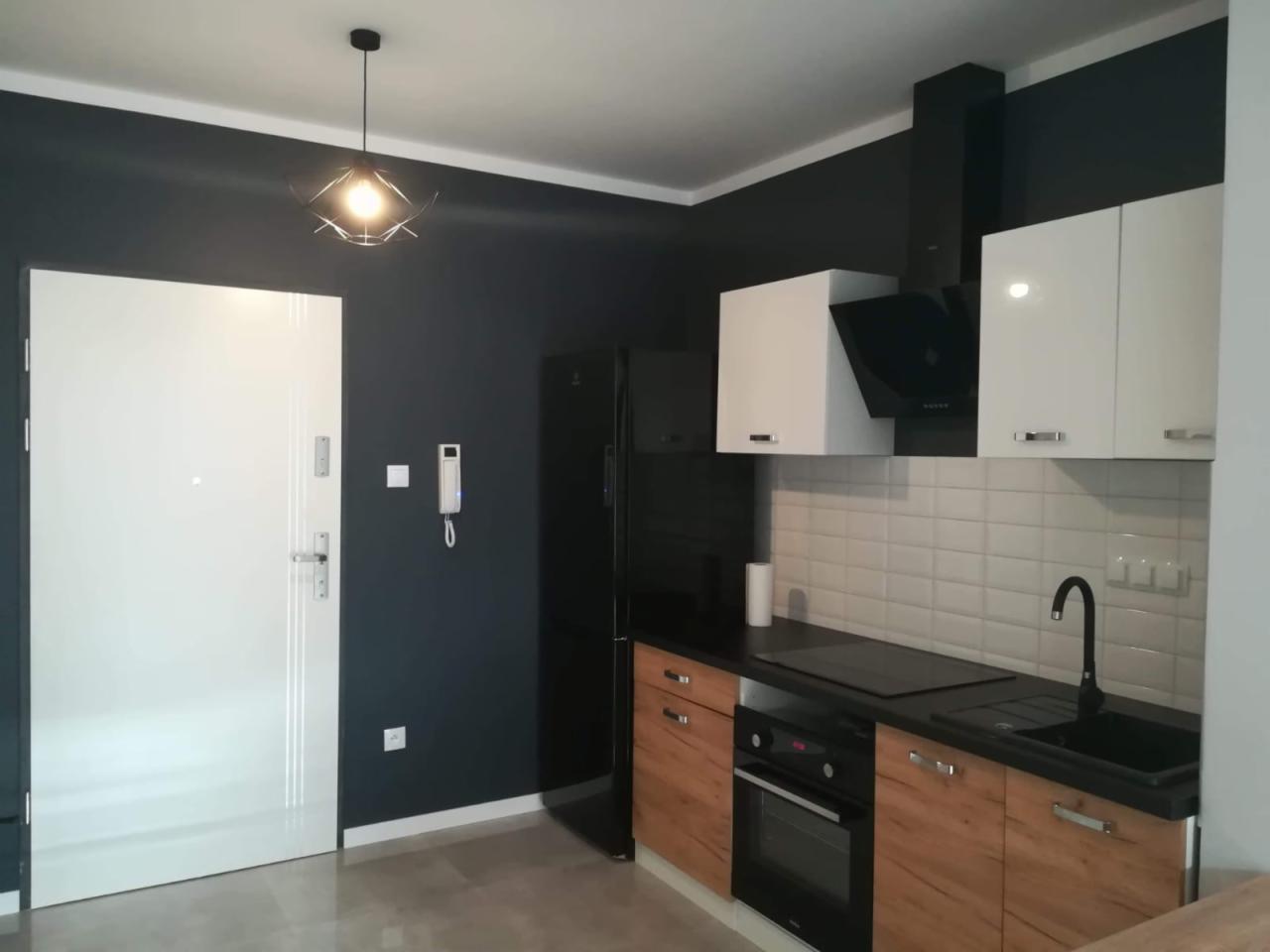 Nowoczesna kuchnia w odcieniach bieli, czerni i drewna, z minimalistyczną lampą wiszącą i białymi płytkami na ścianie.