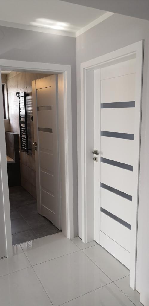 Dwie białe, nowoczesne drzwi wewnętrzne z szarymi wstawkami, widok z korytarza na łazienkę z grzejnikiem drabinkowym i szarą glazurą.