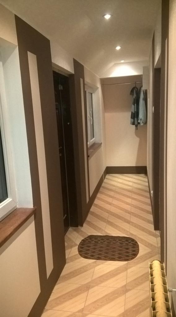 Korytarz z beżowo-brązowymi płytkami ułożonymi w jodełkę, ciemnobrązowe obramowania ścian i drzwi, mały dywanik i grzejnik żeberkowy.