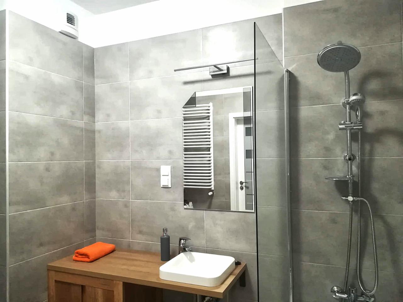 Nowoczesna łazienka z szarymi płytkami, drewnianą szafką pod umywalką, pomarańczowym ręcznikiem, szklanym panelem prysznicowym i chromowaną armaturą.