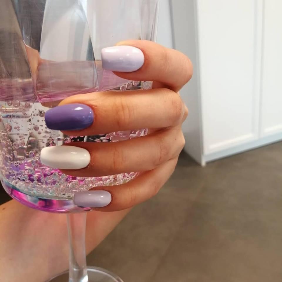 Dłoń z pomalowanymi na różne kolory paznokciami (fioletowy, biały, liliowy) trzyma kieliszek wypełniony wodą z dekoracyjnymi kulkami.