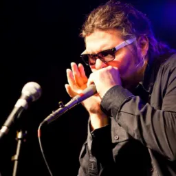 Mężczyzna w okularach gra na harmonijce ustnej podczas występu na scenie, mikrofon blisko ust.