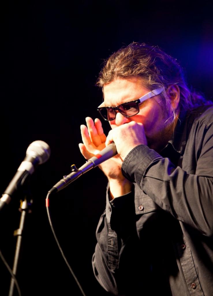 Mężczyzna w okularach gra na harmonijce ustnej podczas występu na scenie, mikrofon blisko ust.