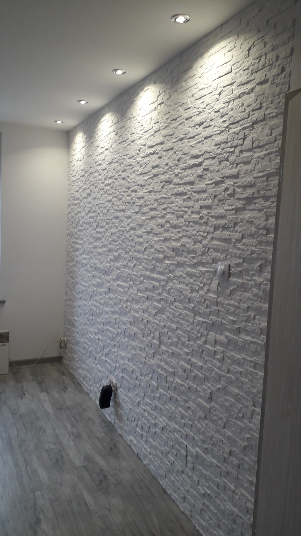Ściana wykończona dekoracyjną, białą okładziną imitującą kamień, z widocznym gniazdkiem elektrycznym i oświetleniem punktowym w suficie, na podłodze panele w odcieniach szarości.