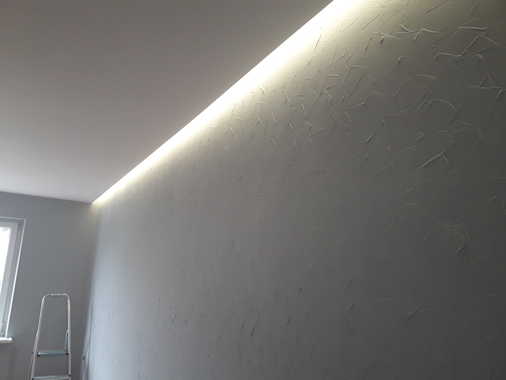 Szara ściana z dekoracyjną fakturą i podświetleniem LED wzdłuż sufitu, widoczna drabina malarska przy oknie.