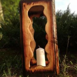 DariaSworowska - Drewniany lampion z wyciętym wzorem, wewnątrz biała świeca, ustawiony na trawie, widoczne ślady opalania drewna.