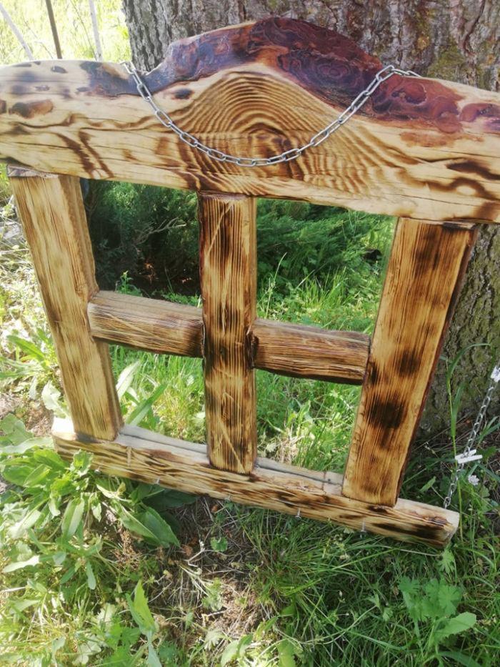 Drewniana konstrukcja z wypalonym wzorem, zawieszona na łańcuchu, w tle trawa i pień drzewa.