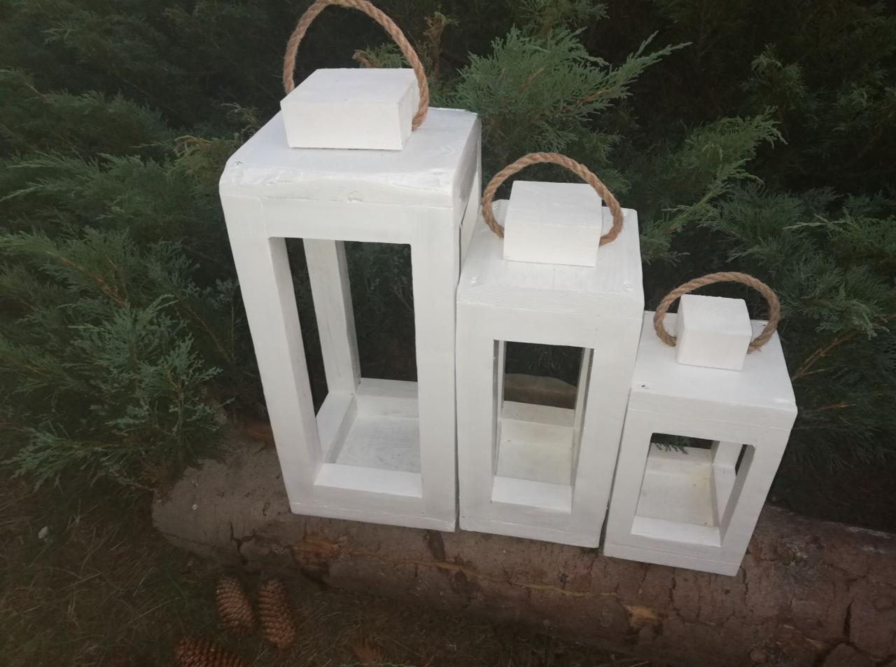 Trzy białe, drewniane lampiony różnej wielkości z jutowymi uchwytami, ustawione na pniu drzewa, na tle zielonych krzewów. Dekoracja ogrodowa.