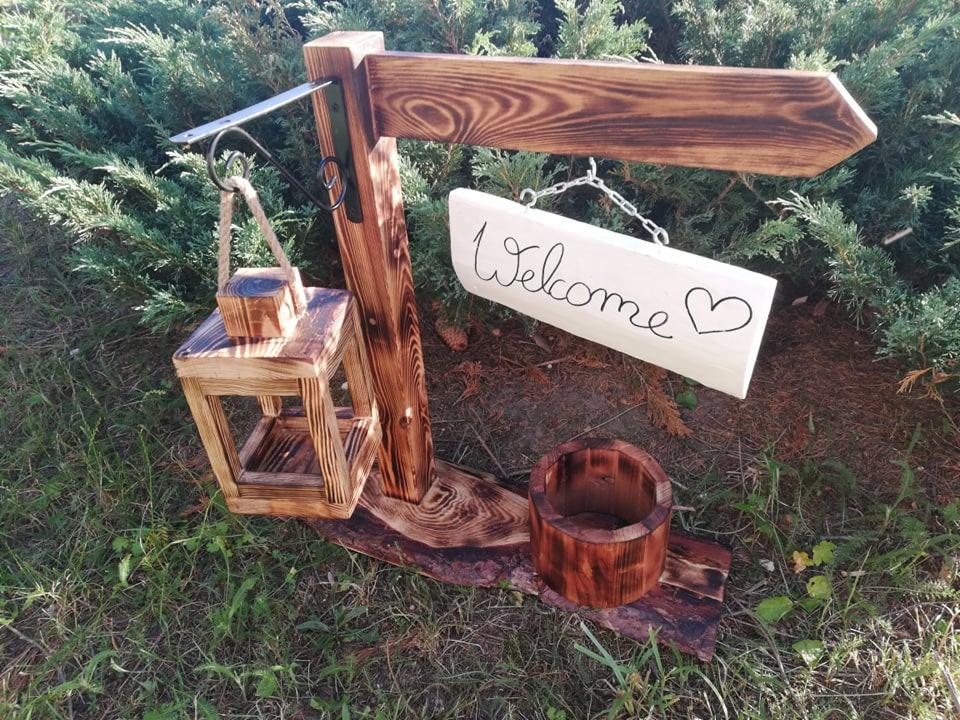 Drewniana dekoracja ogrodowa z drogowskazem, lampionem i napisem 'Welcome' z sercem, umieszczona na trawie przed krzewami.