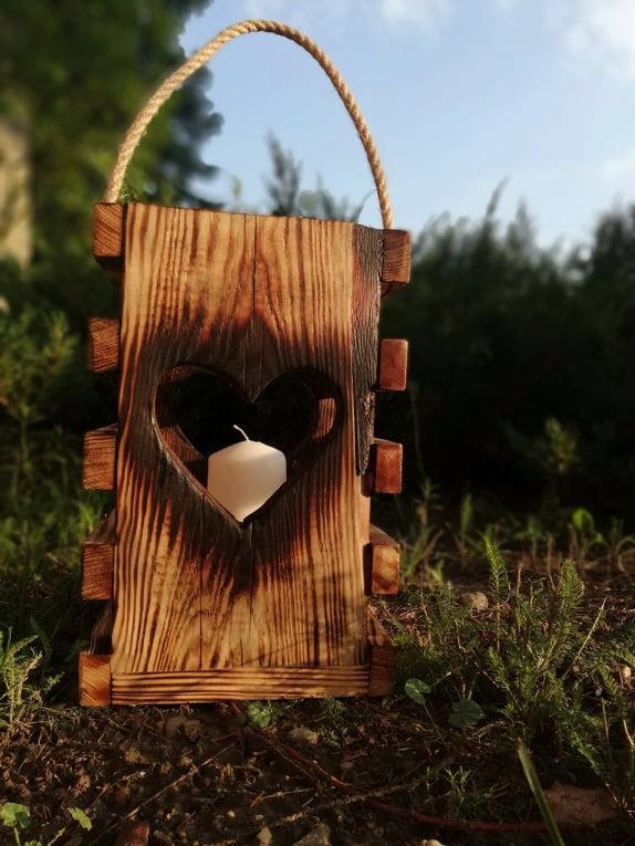 Drewniany lampion z wyciętym sercem i jutowym uchwytem, z białą świecą w środku, na tle zieleni.