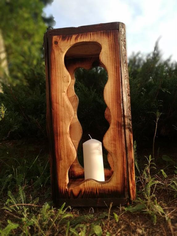 Drewniany lampion z wyciętym wzorem, wewnątrz biała świeca, ustawiony na trawie, widoczne ślady opalania drewna.