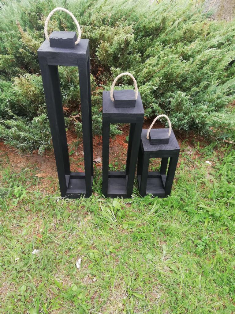 Trzy czarne, drewniane lampiony ogrodowe o różnej wysokości, z jutowymi uchwytami, ustawione na trawie przed zielonymi krzewami.