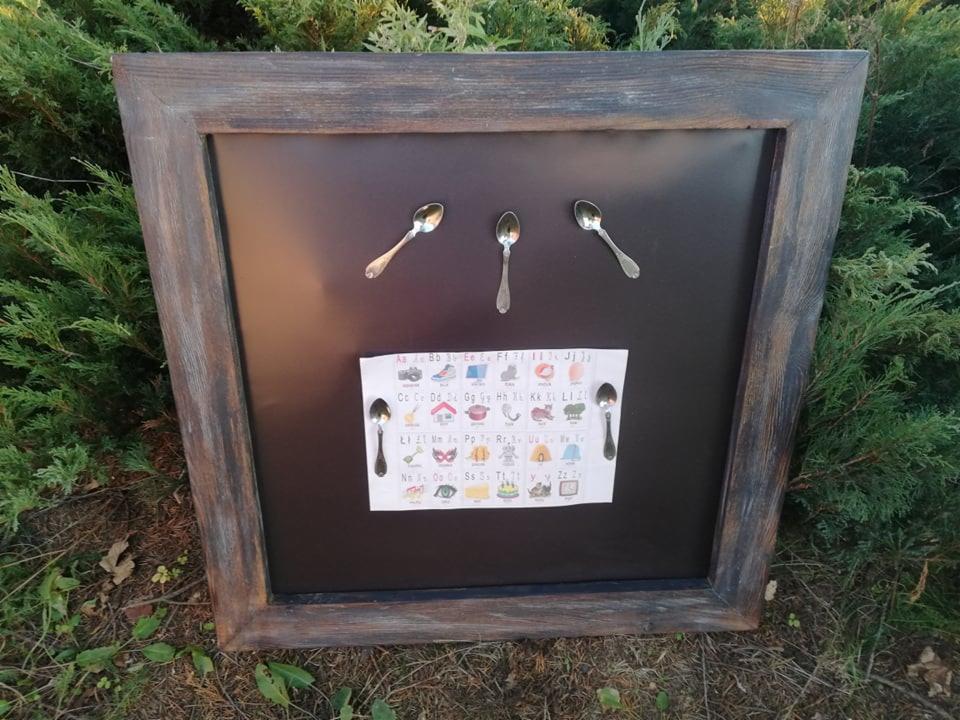 Tablica edukacyjna w drewnianej ramie, z literami alfabetu i obrazkami, przyczepiona do czarnej magnetycznej powierzchni za pomocą czterech srebrnych łyżeczek.